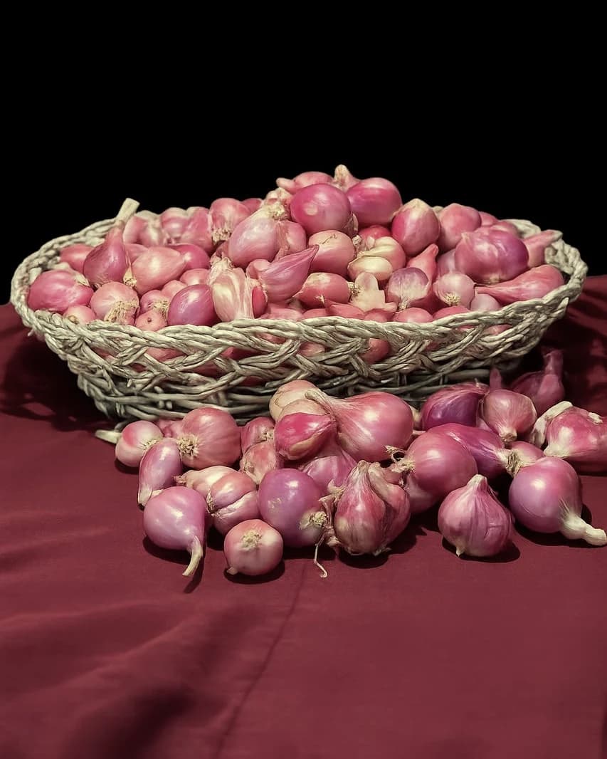 Bawang merah kering BERTARUM menumpuk dalam keranjang anyaman di atas kain merah, sebagian tercecer di depannya.